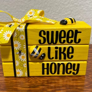 May include: A yellow wooden block set with the words "SWEET LIKE HONEY" in black. The set is decorated with a yellow ribbon with white daisies and bees, and two bee figurines.