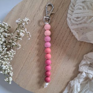 May include: A keychain featuring a vertical row of ten spherical beads in a gradient of pink shades, ranging from light pastel pink at the top to a deep, saturated rose pink at the bottom, attached to a silver-toned metal lobster clasp.