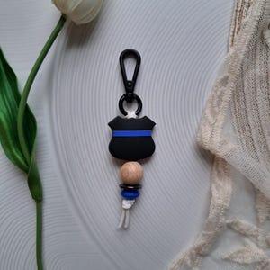May include: Black and blue keychain with a wooden bead and a thin blue stripe. The keychain is shaped like a police badge.