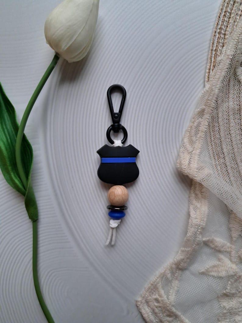 May include: Black and blue keychain with a wooden bead and a thin blue stripe. The keychain is shaped like a police badge.
