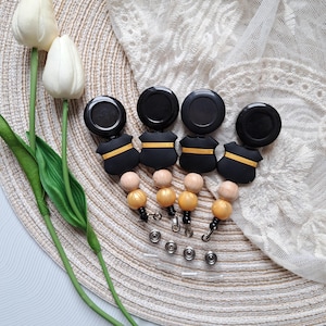 May include: Five black retractable badge reels with gold accents and wooden beads. The reels are on a woven placemat with white lace fabric in the background.