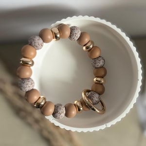 Peut inclure: Un bracelet de perles circulaire composé d'un motif répétitif de perles beige uni, de perles intercalaires dorées métalliques et de perles beige avec un imprimé floral blanc. Un anneau en métal doré est fixé au bracelet, posé dans un bol blanc.