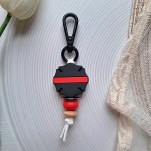 May include: Black and red silicone keychain with a thin red line across the centre. The keychain has a black metal clip and a wooden bead with a red silicone bead.