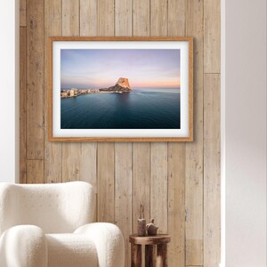 May include: A framed photograph of a rocky island in the ocean with a town in the distance. The sky is a soft purple and pink, and the water is a deep blue. The photo is hanging on a wooden wall.