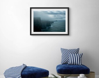 May include: A framed landscape photograph of a coastal cliffside shrouded in mist. The image, in a black frame, depicts dark cliffs meeting the ocean under a cloudy sky. The scene is set against a white wall, with blue furniture and striped pillows.