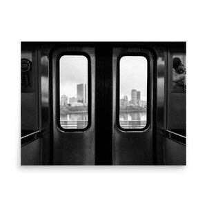 Black and white view of the Boston skyline framed by the doors of an MBTA train, looking out over the Charles River with the Prudential Tower and other city buildings in the distance.