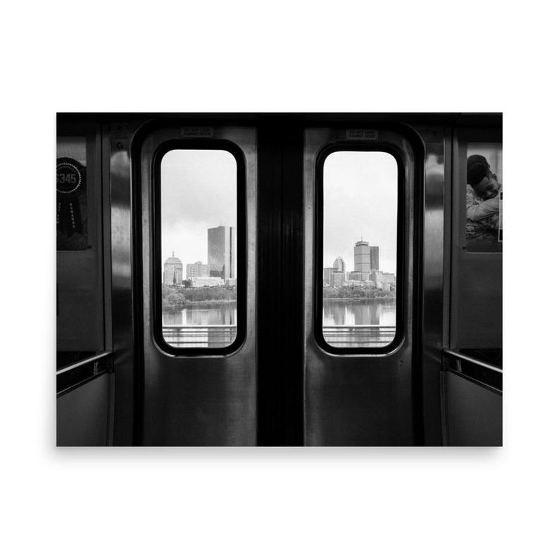 Black and white view of the Boston skyline framed by the doors of an MBTA train, looking out over the Charles River with the Prudential Tower and other city buildings in the distance.