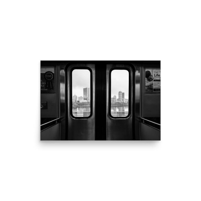 Black and white view of the Boston skyline framed by the doors of an MBTA train, looking out over the Charles River with the Prudential Tower and other city buildings in the distance.