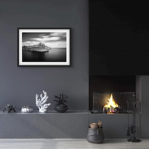 May include: Black and white framed photograph of a coastal scene. The image features a wooden structure on stilts extending into calm water under a cloudy sky. The photograph is displayed on a gray wall above a fireplace.
