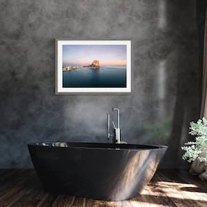 May include: A black bathtub with a silver faucet in front of a gray wall with a framed photograph of a rocky island in the distance.