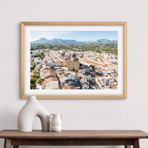 May include: An aerial view of a small town in Spain with a white church in the center. The town is surrounded by mountains and green trees.