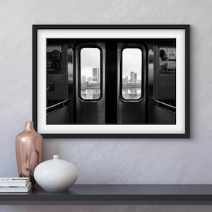 Pode incluir: Fotografia emoldurada a preto e branco do interior de um vagão de metro, com vista para o horizonte da cidade através das portas abertas. A fotografia é exibida numa mesa cinzenta escura, com vasos decorativos e livros.