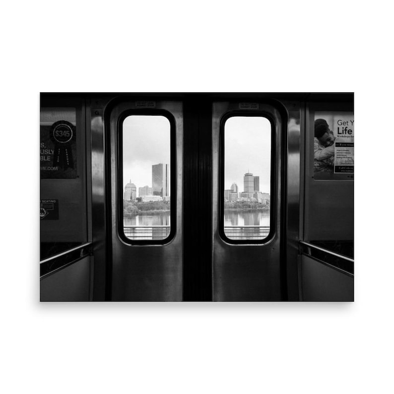 Black and white view of the Boston skyline framed by the doors of an MBTA train, looking out over the Charles River with the Prudential Tower and other city buildings in the distance.