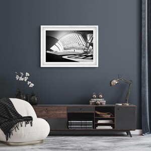 May include: A black and white photograph of a modern building with a curved roof and a large open space. The photo is framed in a white frame and hanging on a dark blue wall.