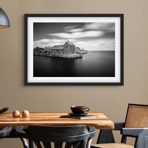 May include: Black and white photograph of a wooden building on a pier with a cloudy sky and calm water in the background.