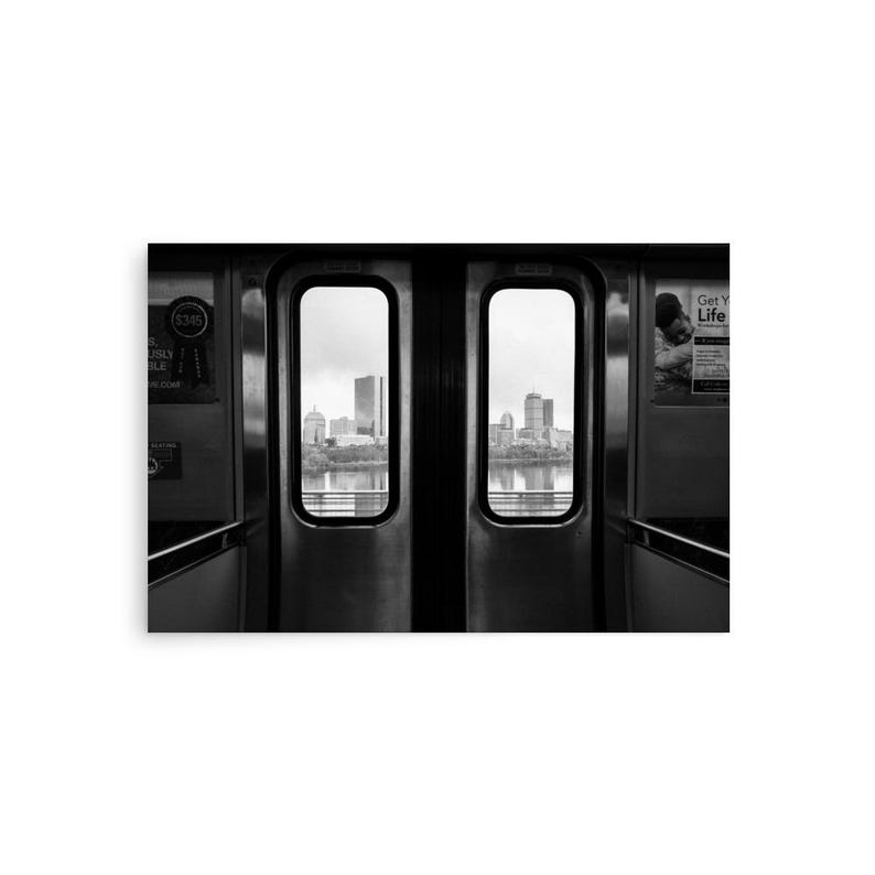 Black and white view of the Boston skyline framed by the doors of an MBTA train, looking out over the Charles River with the Prudential Tower and other city buildings in the distance.