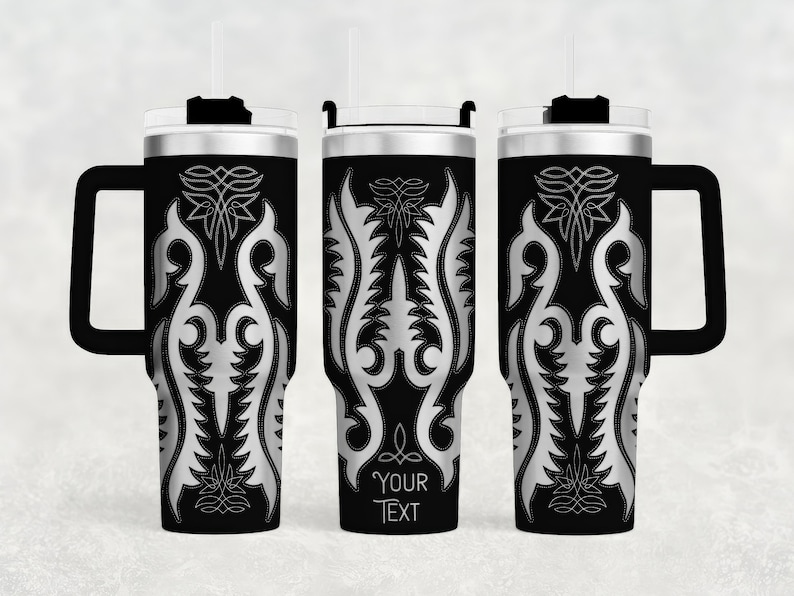 May include: Black insulated tumbler with a silver ornamental design. The tumbler has a handle, a lid, and a straw. The text "YOUR TEXT" is printed on the tumbler. The design is symmetrical and features floral and geometric elements.