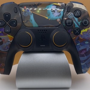 May include: Black and dark blue PlayStation 5 controller with a Halloween-themed design. The controller features a variety of cartoon characters and symbols. The controller is resting on a silver stand.