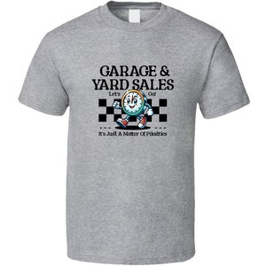Puede incluir: Camiseta gris con un personaje de dibujos animados en forma de reloj corriendo sobre una línea de meta a cuadros. El texto dice "Garage & Yard Sales" con las palabras "Let's Go!" encima del reloj y "It's Just A Matter Of Priorities" debajo.