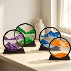 May include: Four circular sand art displays with black frames and stands. Two frames contain purple and green sand, one contains blue sand, and one contains orange sand. Each display features layered sand forming mountain-like landscapes.