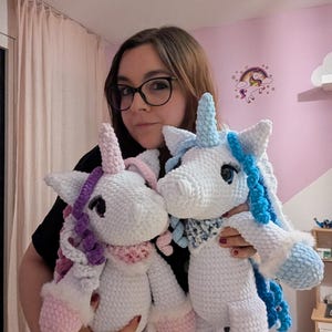 May include: Two handmade crochet unicorn plush toys. The unicorns are white with pink, purple, and blue accents. They have fluffy manes, horns, and hooves. Each unicorn has a scarf around its neck.