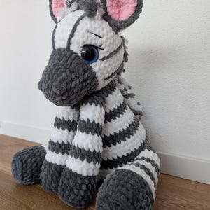 Könnte beinhalten: Ein Plüschzebra aus weichem, strukturiertem Garn mit abwechselnden horizontalen Streifen in Weiß und Dunkelgrau. Das Stofftier hat große, dunkle Augen mit blauen Akzenten, abgerundete Ohren mit rosa Innendetails und eine graue Schnauze.