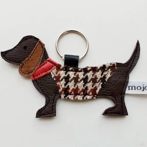 May include: Brown leather dachshund keychain with a red and brown patterned vest and a black key ring. The keychain has a white tag with the word "mojose" printed on it.