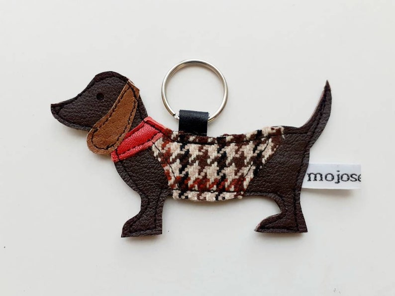May include: Brown leather dachshund keychain with a red and brown patterned vest and a black key ring. The keychain has a white tag with the word "mojose" printed on it.
