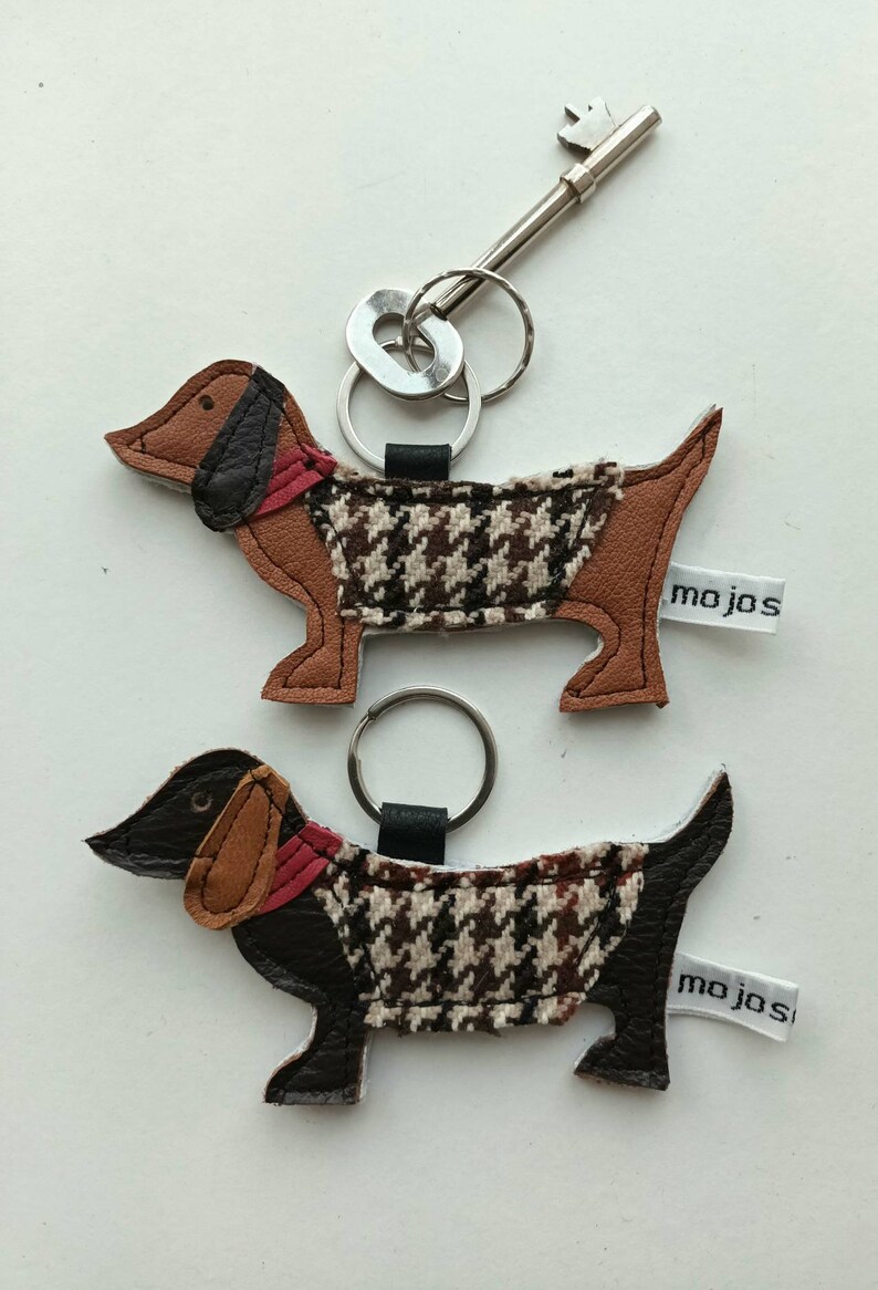 May include: Two brown and black dachshund dog keychains with red collars and a houndstooth pattern. Each keychain has a silver key ring and a white tag with the text "mojos".