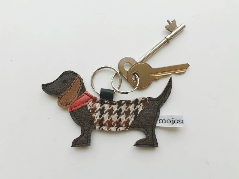 May include: A brown dachshund-shaped leather keychain with a red and brown patterned fabric vest. The keychain has a silver key ring and is attached to a silver key.