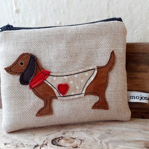 Peut inclure: Une pochette zippée en lin beige avec une applique de dachshund marron portant une écharpe rouge et une robe à pois blancs avec un cœur rouge. La pochette a une fermeture éclair noire et une étiquette blanche qui dit "mojos".