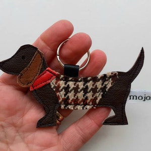 May include: Brown leather dachshund keychain with a red and brown patterned vest and a silver key ring. The keychain is labeled "mojose".