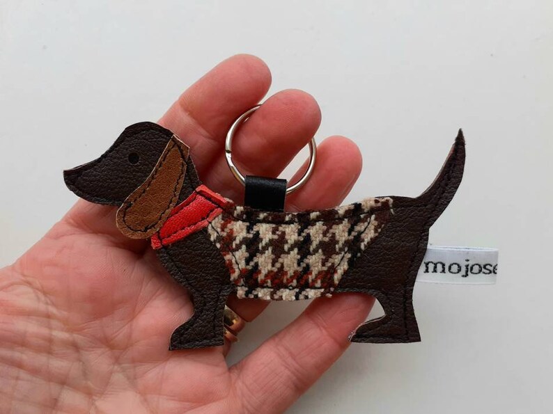 May include: Brown leather dachshund keychain with a red and brown patterned vest and a silver key ring. The keychain is labeled "mojose".