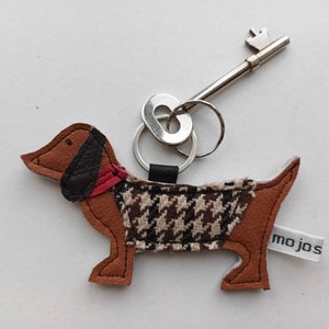 May include: A brown leather dachshund keychain with a black and white houndstooth pattern on the body and a red collar. The keychain has a silver key ring and a silver key with a decorative top. The keychain has a white tag with the word "mojos" printed on it.