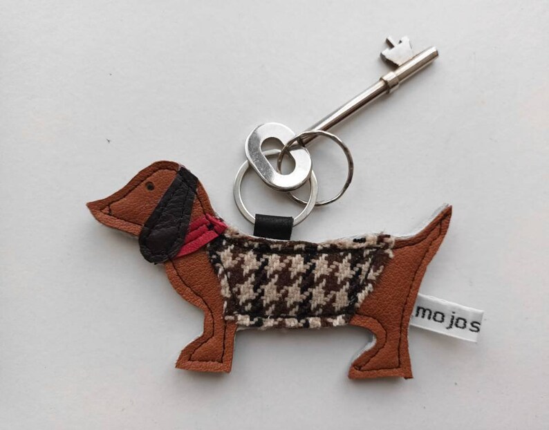 May include: A brown leather dachshund keychain with a black and white houndstooth pattern on the body and a red collar. The keychain has a silver key ring and a silver key with a decorative top. The keychain has a white tag with the word "mojos" printed on it.