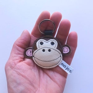 Peut inclure: Un porte-clés en cuir marron en forme de singe avec des oreilles roses et un visage blanc. Le porte-clés a un anneau de clé argenté et une étiquette blanche avec le mot "mojos" imprimé dessus.
