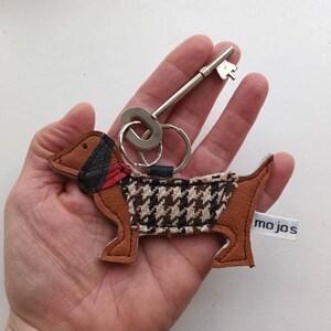 May include: A brown and black dachshund-shaped leather keychain with a houndstooth pattern and a red collar. The keychain is attached to a silver key ring and a silver key. The keychain has a white tag with the text "mojos" on it.