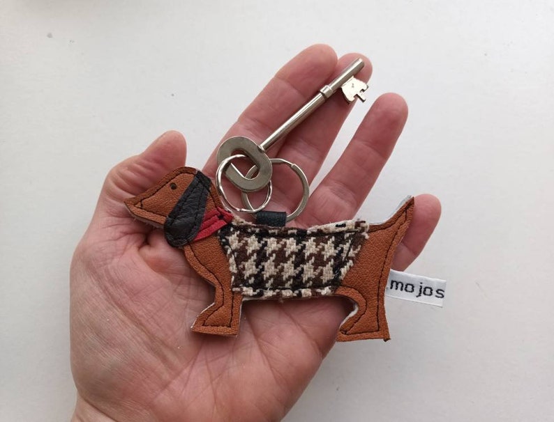 May include: A brown and black dachshund-shaped leather keychain with a houndstooth pattern and a red collar. The keychain is attached to a silver key ring and a silver key. The keychain has a white tag with the text "mojos" on it.
