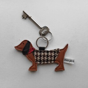 May include: A brown and black dachshund-shaped leather keychain with a black and white houndstooth pattern on the body. The keychain has a red scarf and a silver key ring attached.