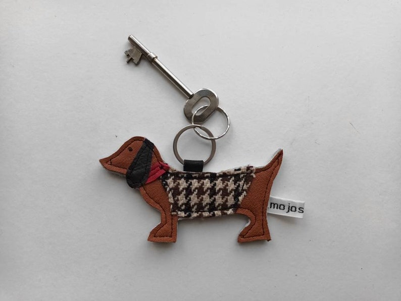 May include: A brown and black dachshund-shaped leather keychain with a black and white houndstooth pattern on the body. The keychain has a red scarf and a silver key ring attached.