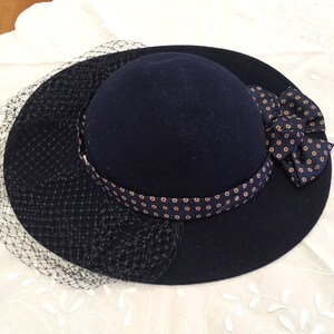 May include: A navy blue hat with a rounded crown and wide brim. The hat features a patterned band and a bow on the side. A black netting veil drapes over the brim. The hat is likely made of felt or a similar material.