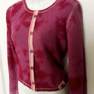 May include: A pink tie-dye cardigan with a white stripe down the front and colorful buttons. The cardigan has long sleeves and a cropped length.