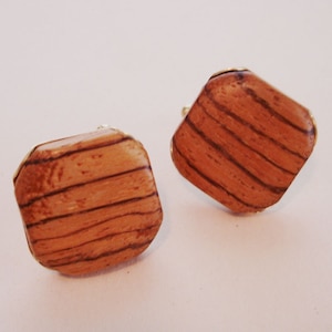 May include: A pair of square wooden cufflinks with a wood grain pattern. The cufflinks are a light brown color and have a gold-colored metal trim.