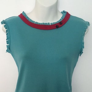 May include: Teal sleeveless shirt with a red ruffled neckline and two black buttons. The shirt has a frayed edge around the neckline and armholes.