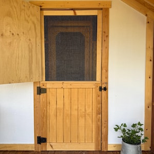 Puede incluir: Una puerta de madera con una pantalla y una pequeña puerta de madera está abierta. La puerta es de madera clara con tablones verticales. La puerta mosquitera está enmarcada en madera. Un cubo de metal galvanizado con una planta verde está a la derecha.