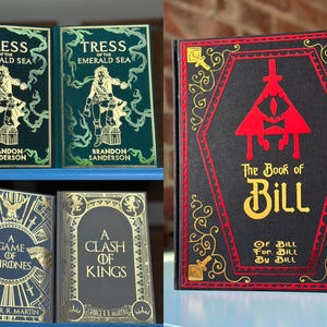 Op de afbeelding: Afbeelding van verschillende boeken met unieke covers. Twee exemplaren van "Tress of the Emerald Sea" van Brandon Sanderson staan op de bovenste plank. Hieronder staan "A Game of Thrones" en "A Clash of Kings" van George R.R. Martin. De rechterkant toont "The Book of Bill".