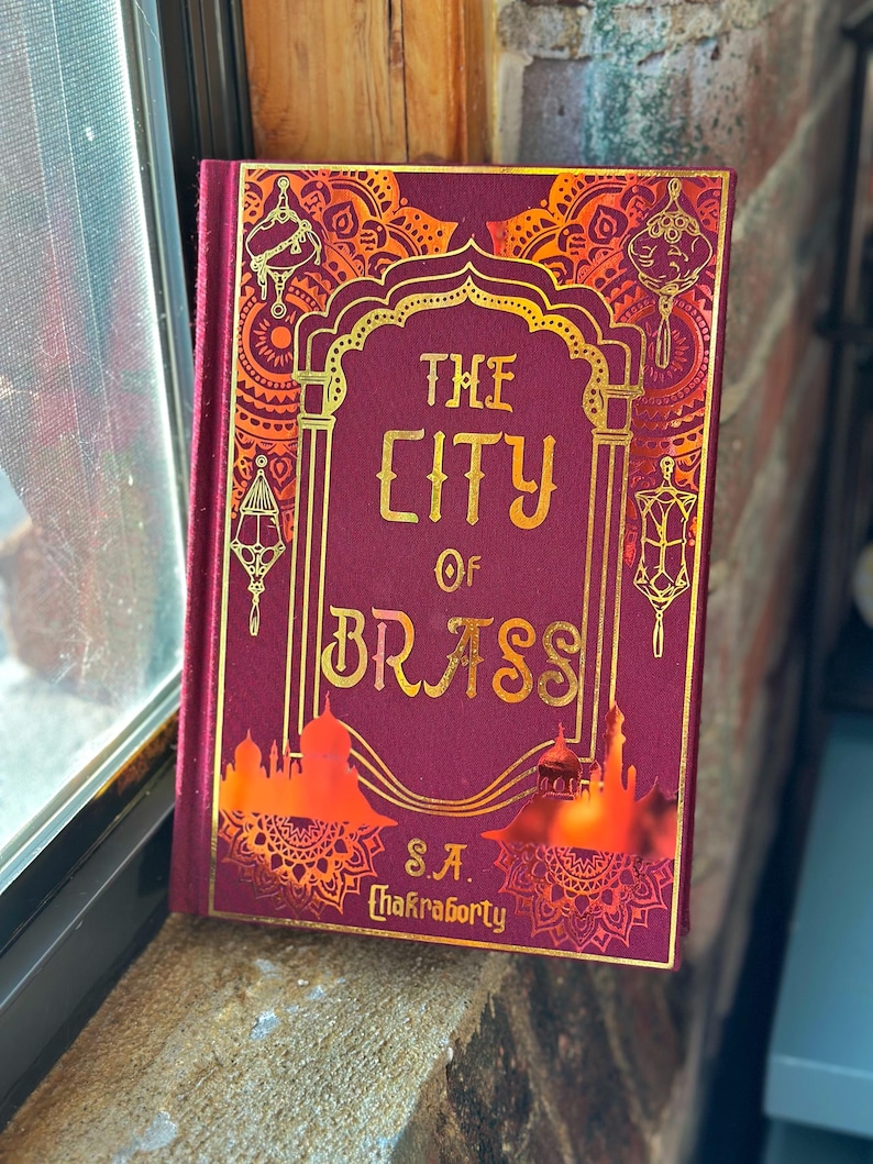 Op de afbeelding: Een hardcover boek met de titel "The City of Brass" van S.A. Chakraborty. Het boek heeft een bordeauxrode kaft met gouden letters en decoratieve accenten. Het omslagontwerp bevat sierlijke patronen en architecturale silhouetten.