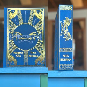 以下が含まれることがあります： 金色の文字とイラストが施された、青いハードカバーの書籍「Dragonlance Chronicles」。 本は青い棚に置かれ、表紙と背表紙が示されています。
