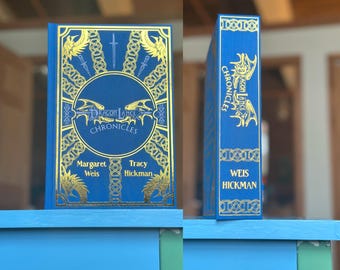 Könnte beinhalten: Ein blaues Hardcover-Buch mit dem Titel "Dragonlance Chronicles" mit goldfarbenen Buchstaben und Illustrationen. Das Buch wird auf einem blauen Regal präsentiert und zeigt den Einband und den Buchrücken.