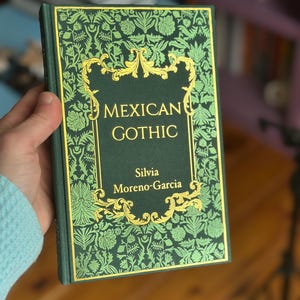 Op de afbeelding: Een hardcover boek getiteld "MEXICAN GOTHIC" van Silvia Moreno-Garcia. Het boek heeft een donkergroene kaft met een gouden bloemenontwerp en gouden letters. Het boek wordt in een hand gehouden.